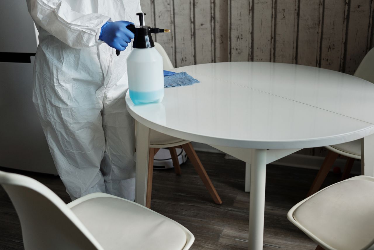 A person in a protective suit disinfecting a white table indoors using a sprayer.